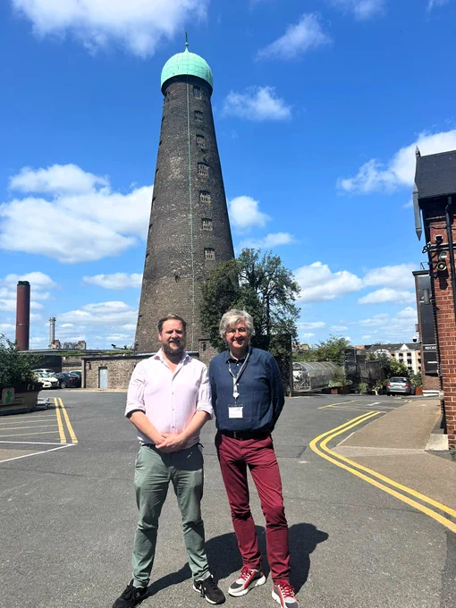 Two people, Patrick Costello TD & Fiach Mac Conghail, CEO of The Digital Hub, are standing in front of a tall tower called St. Patrick's Tower. The tower is topped with a green copper dome and the sky is blue. Both Patrick & Fiach are standing in a relaxed pose and smiling.