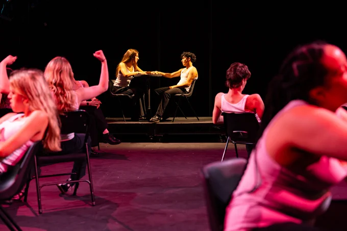 Two people sitting on chairs facing each other, engaged in an animated conversation with hands gesturing, on a dimly lit stage. In the foreground, blurred figures are seated on chairs in various poses