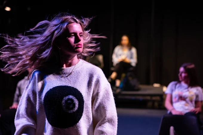 A person with flowing hair wearing a sweater with an 8-ball design is dynamically moving on a stage, with two other individuals seated in the background, slightly out of focus, in a black box theatre space