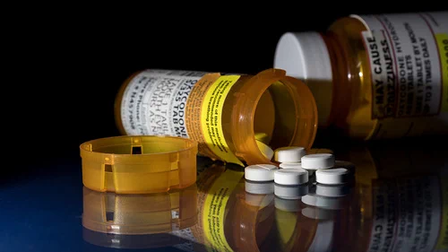Several prescription medication bottles lying on their sides with their caps open and white pills spilled out on a reflective surface.