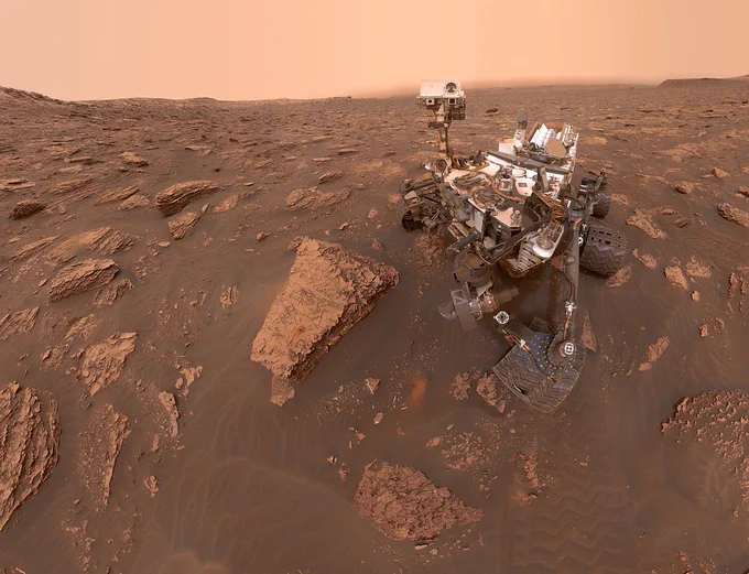 The Curiosity rover is shown in a selfie in a rocky area of Mars. The image is a rust color and the rover is lightly covered in Martian dust. It is slightly off center to the right and is staring at the camera on its robotic arm that took the photo. Next to the left side of the rover is a large reddish rock. In the background are small hills and haze along the horizon.