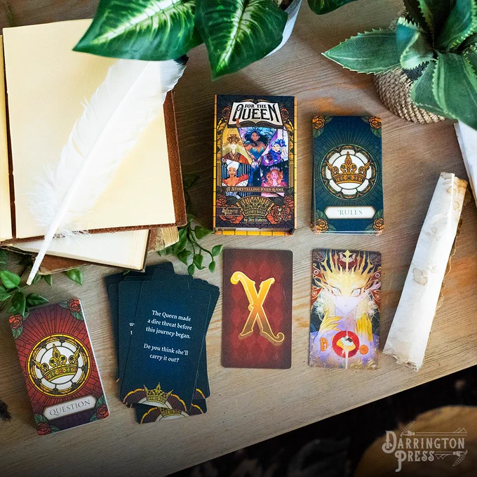 Photography of the storytelling card game Queen by Midnight on a table, with beautiful Queen illustration cards, prompt cards, and an X card in addition to plants, a journal, and a scroll nearby.