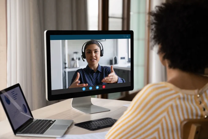 A person is engaging in a video call with a colleague who is gesturing positively on the screen. They are seated in a home office environment, with a laptop and documents visible on the desk, emphasizing a professional remote work setting.