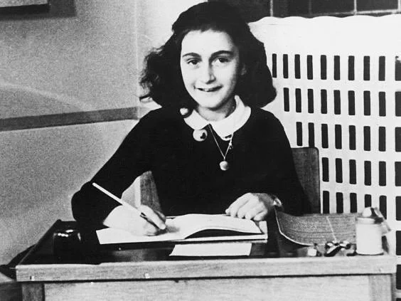 Black and white historical photo of Anne Frank sitting at a desk writing in a notebook. She is wearing a dark blouse with a white collar and has short hair. The background features a simple paneled wall.