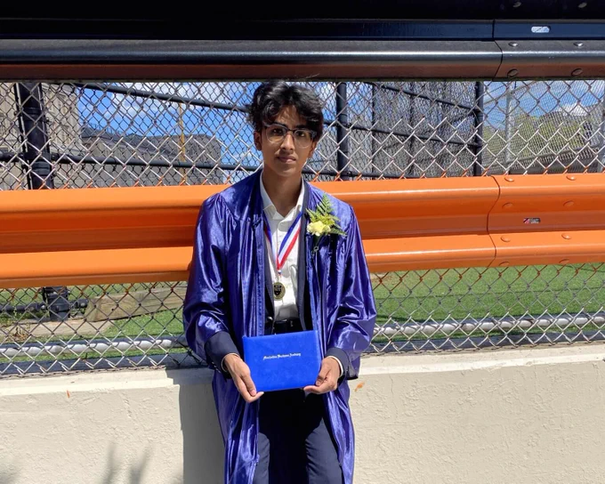 A student poses in a blue graduation gown while holding a diploma. 