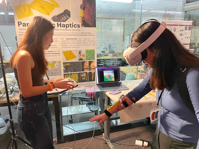 A female researcher is demoing their new haptic device. A user is wearing the device and a VR headset.