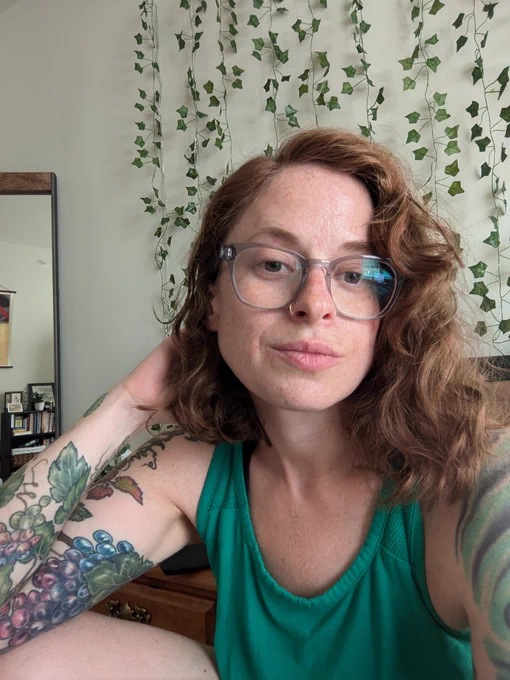 photo of me, red headed bespectacled dummy, sitting in front of a wall decorated with fake ivy vines, very subtly showing off my new tattoo sleeve by propping my arm up behind my head.