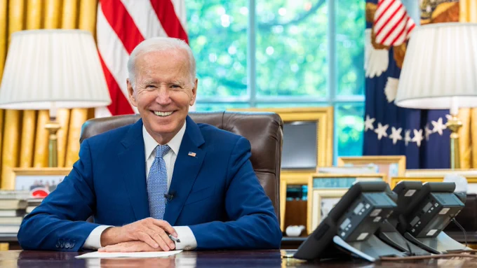 President Joe Biden sitting at a desk in the Oval Office, smiling, with the American flag in the background.
