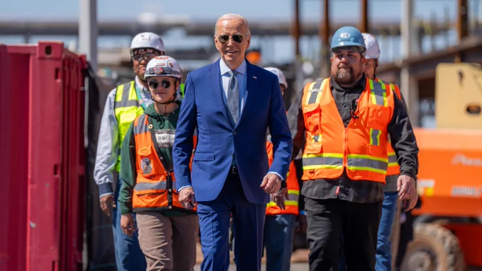 President Joe Biden walks alongside construction workers wearing safety vests and helmets at an industrial site.