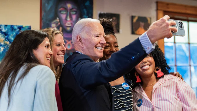 President Joe Biden takes a selfie with two supporters, with colorful artwork visible in the background.