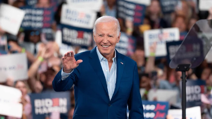 Joe Biden is pictured waving and smiling broadly at a campaign event. He is dressed in a dark blue suit and a flag pin on his lapel, walking in front of a crowd with signs, some reading "Biden Harris."