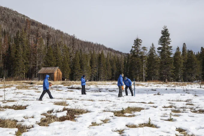 Managers and engineers in winter clothes conduct the first snow survey of the year in the Sierra Nevada. They are in a field with a small layer of snow on it with mountains in the background.