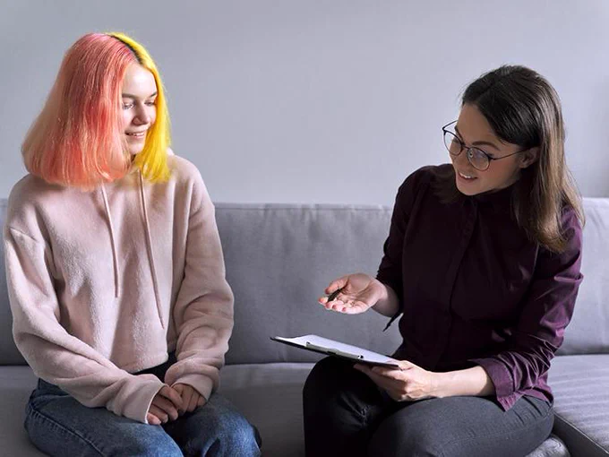 Two young women are having a discussion while seated on a couch; one holding a clipboard and pointing at it, and the other listening attentively.