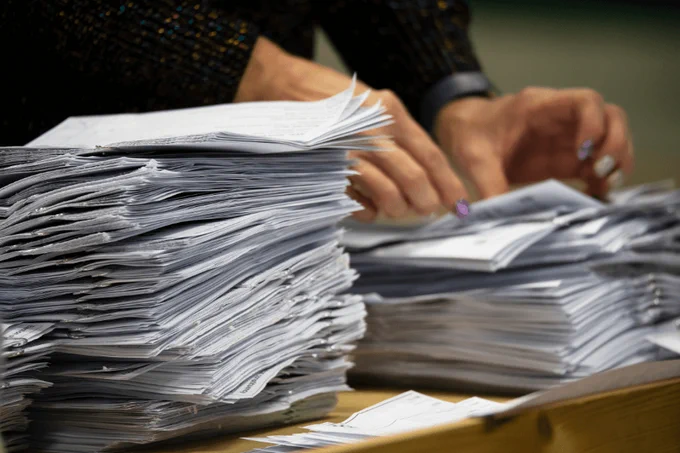 Multiple stacks of papers on a wooden table, with a person moving some of the papers in the background.
