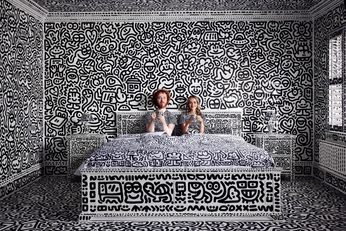 A man and woman sit up in a bed looking at the camera. The room they are in is painted white and covered in doodles in black. This includes the bed, the bedding and the walls.