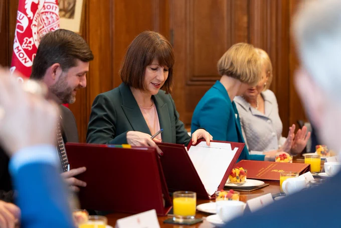 The Chancellor is pictured seated while wearing a green jacket. She is looking at her notes in a red folder. To her right and left are officials and senior leaders from the pensions sector. Behind her is wooden panelling and a HM Treasury flag.