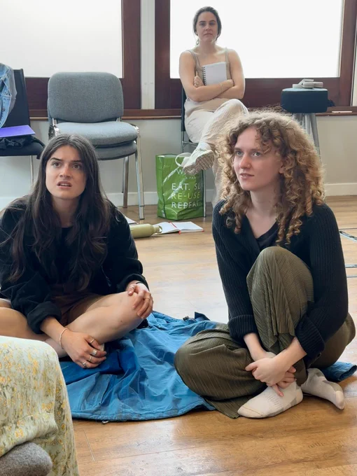 Mimi and Ellen sit on the floor looking up towards somebody out of shot. Chan sits on a chair in the background. They are all white women.