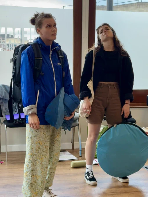 Maddie and Mimi stand looking frustrated, in character. They are both white women. Mimi holds a pop up tent bag.
