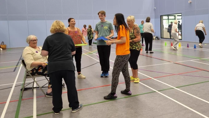 A group of people stand in a circle in a sports hall. There are people in the background taking part in games.