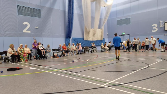 A picture of a sports hall. Lots of people sat on chairs around the edge, waiting to take part in activities.