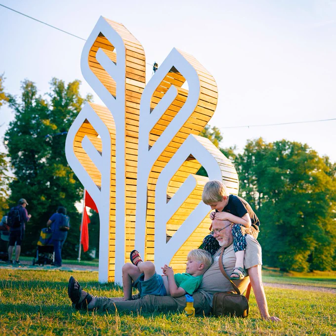 Two small children climbing on their grandpa in front of a large wood sculpture