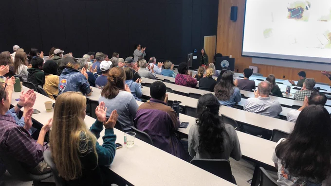 A lecture hall full of people applauding for Dr. Adrienne Correa, who is standing in front of the podium, having just completed a lecture.