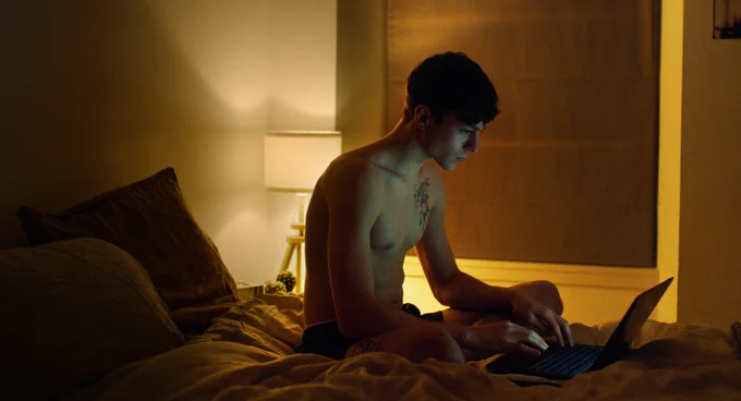 A person sitting on a bed in a dimly lit room, using a laptop