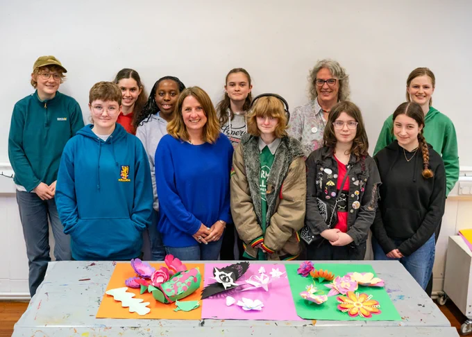 Nine teenagers and two tutors stand behind a table full of paper flowers, smiling at the camera