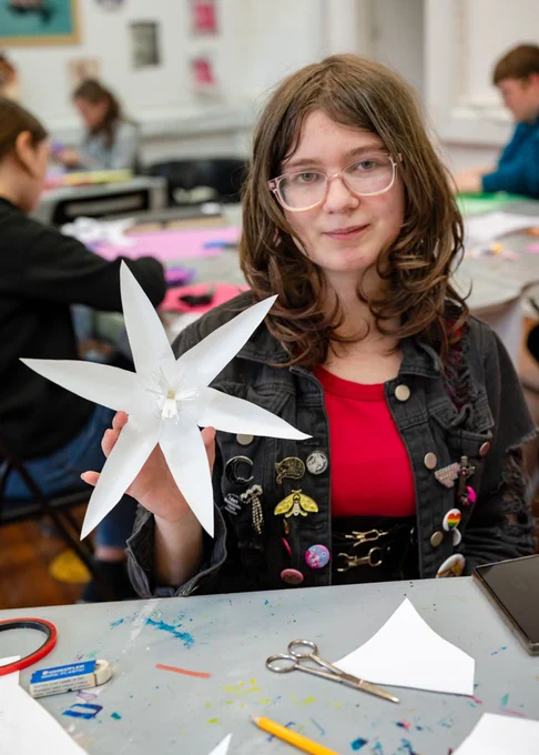 A young person wearing a red top, jacket with badges and pink glasses holds up a white paper flower they made