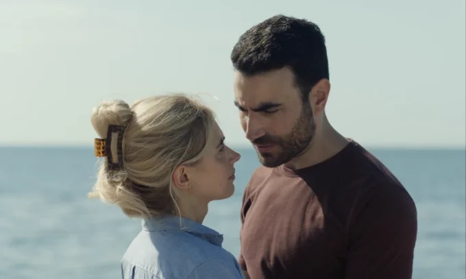 A film still from All of You. A man and a woman stand close together, gazing at each other, in front of a body of water. He has dark hair and a beard, and is wearing a brown shirt. She has long blonde hair up in a clip, and is wearing a light blue button-down.
