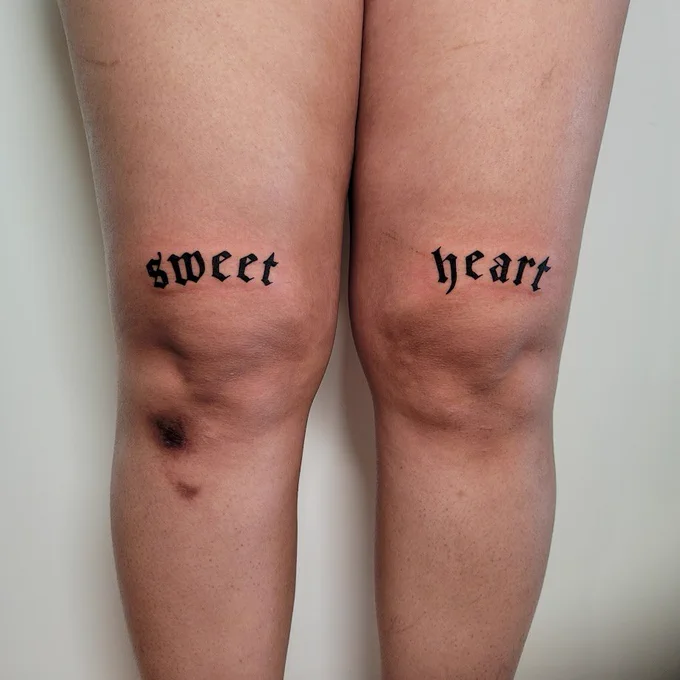 fresh tattoo on the upper knee of a person. there is arched gothic text above each knee cap that reads "sweet" and "heart" and is filled in with black ink.
