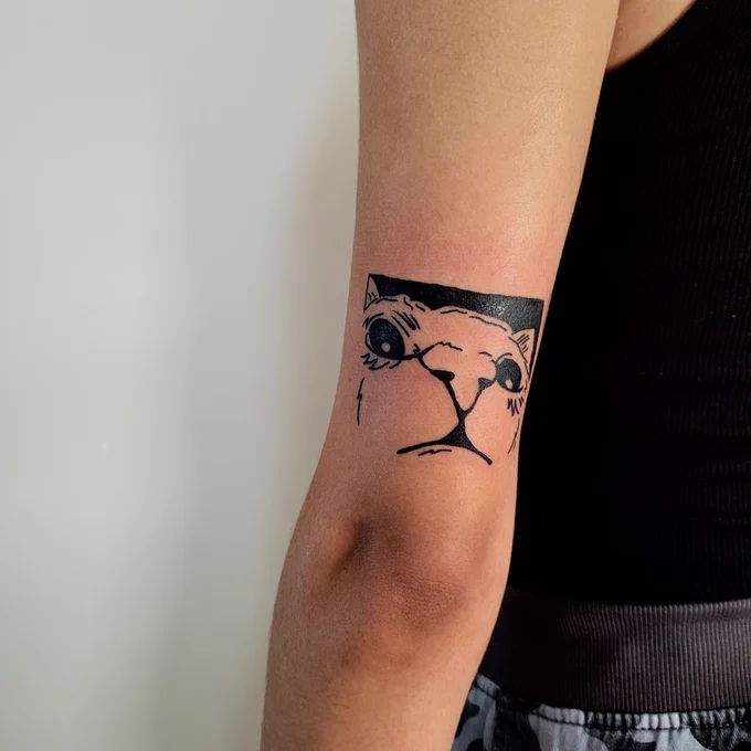 photo of a fresh tattoo on the back of the arm above the elbow on a person. the tattoo is a blackwork style rendering of a cat looking somewhat confused with a 0.5-zoom angle. this makes the nose the star and centre of the piece. the cat has a squared off black background at the top of the piece to shape it which rests nicely on the curve of the back of the arm.