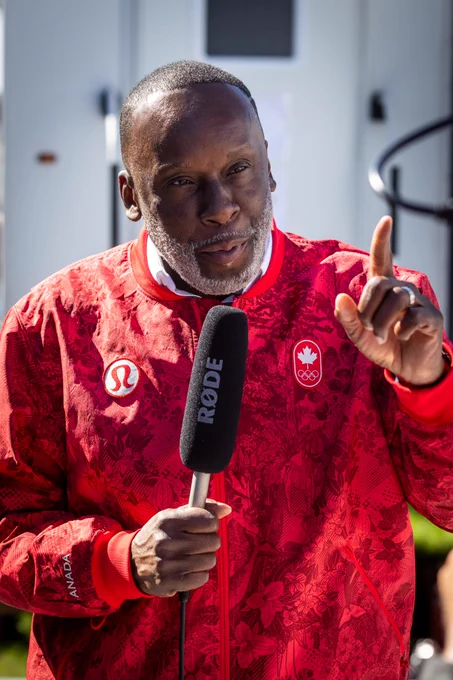 Bruny Surin, Team Canada Chef de Mission, being interviewed, holding a microphone, and raising one finger. He is wearing a red Team Canada jacket.