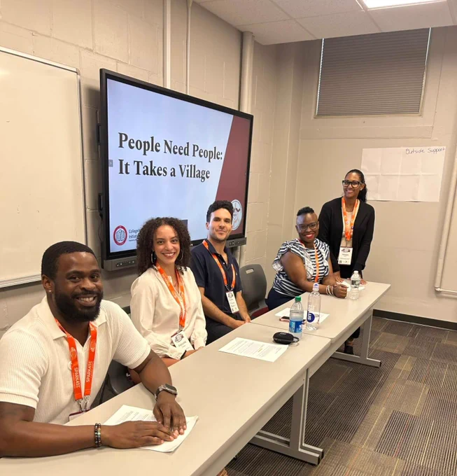 Staff sit at a table with a presentation behind them that says, “People Need People: It Takes a Village.”