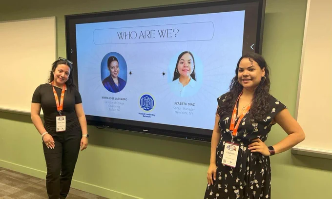 Two women smile and pose in front of a presentation screen that says “Who Are We?” with their headshots. 