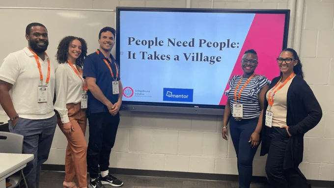Staff smile and pose in front of a presentation screen that says, “People Need People: It Takes a Village.”