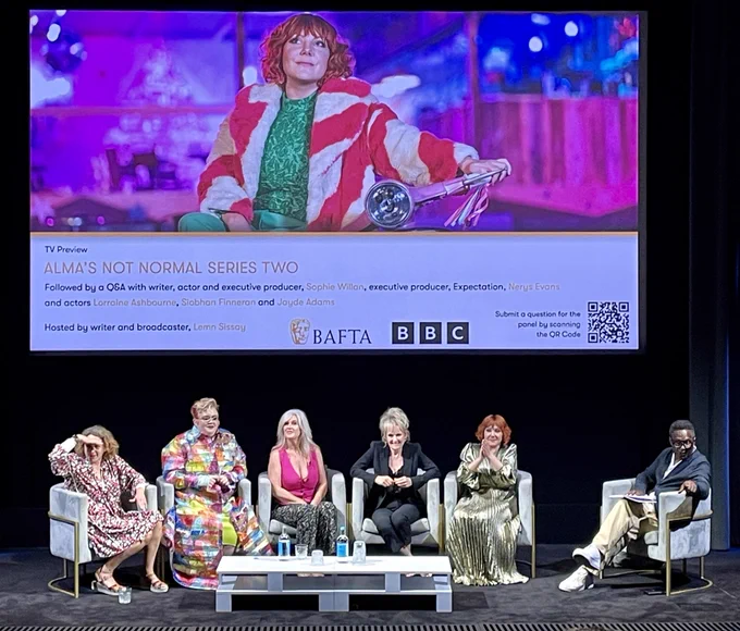 BAFTA stage. Screen shows  Sophie Willan as Alma in a production shot for Series 2 of Alma’s Not Normal. Main cast seated on stage beneath. Sophie, wearing a long gold dress, has hands clasped in a ‘thank you’ gesture.