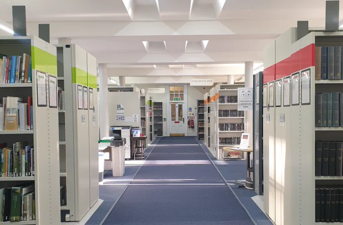 Colour photograph of the interior of a library. Either side of a long, carpeted walkway are rows of moveable shelving with colour-coded bands at the top of each unit. At the far end of the walkway is a door.