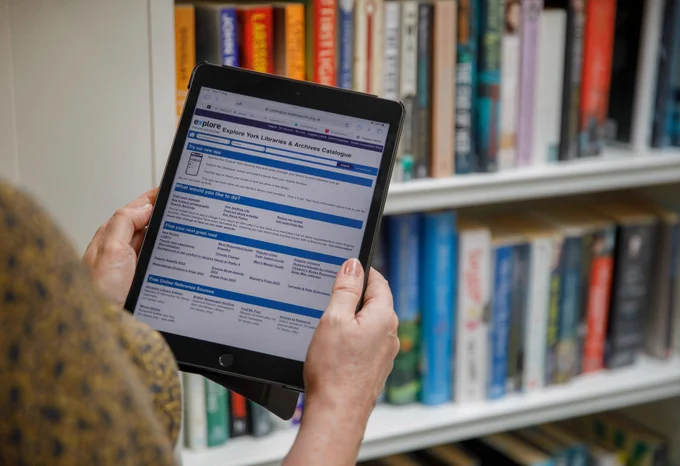 A photograph of hands holding an electronic tablet, on the Catalogue screen, with a background of bookcases.