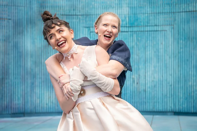 Letty Thomas wearing a blue dress hugging Christina Tedders, wearing a peach coloured dress, from behind.