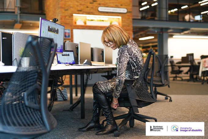 Professional adjusting their desk chair in an office. The Business Disability Forum 'Changing the image of disability' logo is in the lower right corner of the image. 