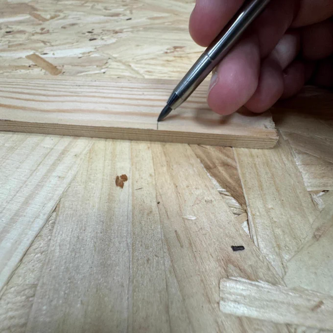 A person marking a measurement on a wooden board with a pen, demonstrating a step in woodworking or carpentry on a wooden workbench.