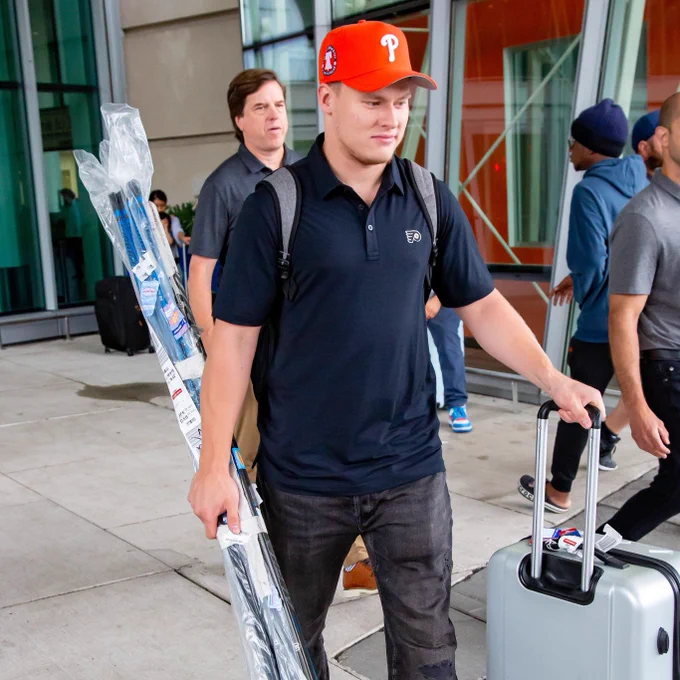 Matvei Michkov standing outside the airport holding his sticks and suitcase. He is wearing an orange Phillies hat and a black Philadelphia Flyers shirt.
