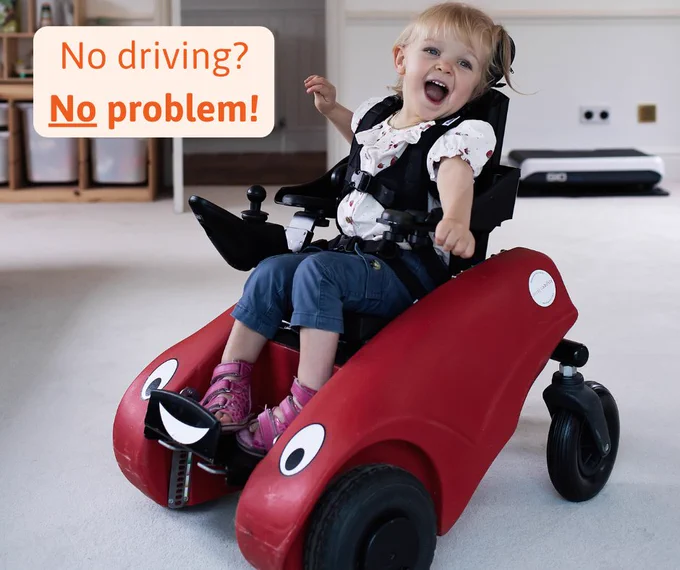 A child looks happy as can be in their Wizzybug powered wheelchair while at home with text: No driving? No problem!