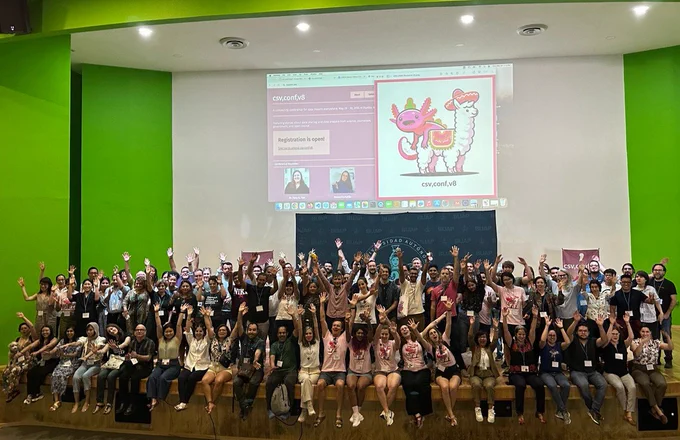Group photo of csv,conf attendees, the background is a colorful csv,conf banner