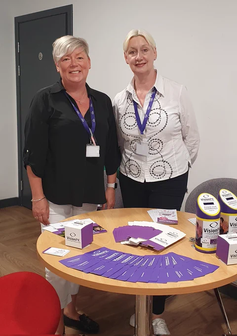 VisionPK services manager and fundraising manager smiling standing behind a desk containing information 