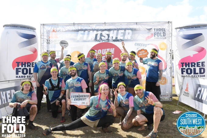 A group of employees from Auroch Digital are grinning at the camera while covered in mud after completing Tough Mudder.