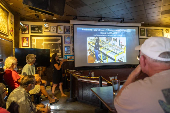 Tessa Hill points at a screen displaying a presentation while a group of people gather around, listening