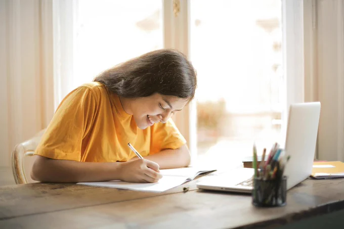 Picture of a person writing in a notebook with a laptop in front of them.