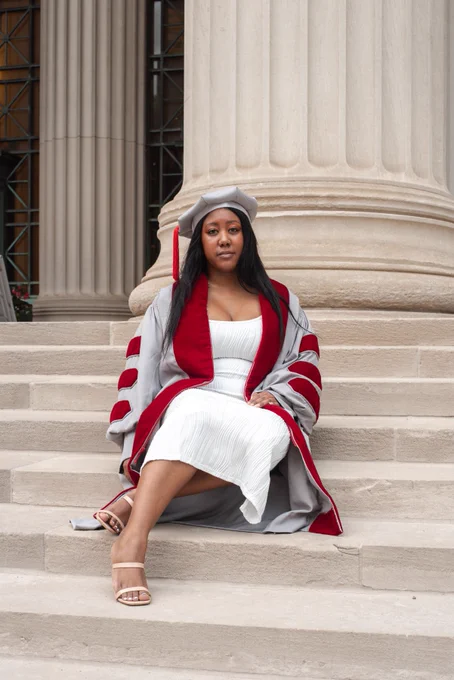 I'm sitting on steps with my left leg crossed behind the right. I'm wearing a white dress, burgundy and gray phd gown and gray cap. I'm wearing tan sandals. There are gray pillars behind me.